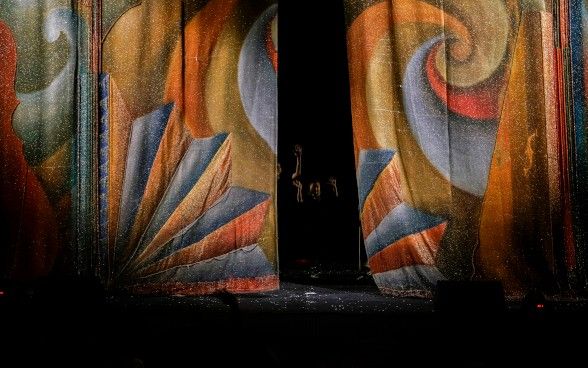 Un sipario semiaperto. Dietro, nel buio, si scorgono alcune persone.