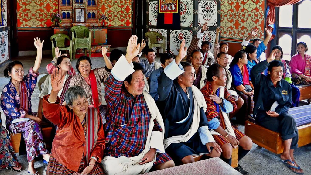 Cittadine e cittadini di tutte le età seduti su delle panche in una comunità rurale del Bangladesh votano per alzata di mano nel processo di pianificazione della governance locale.
