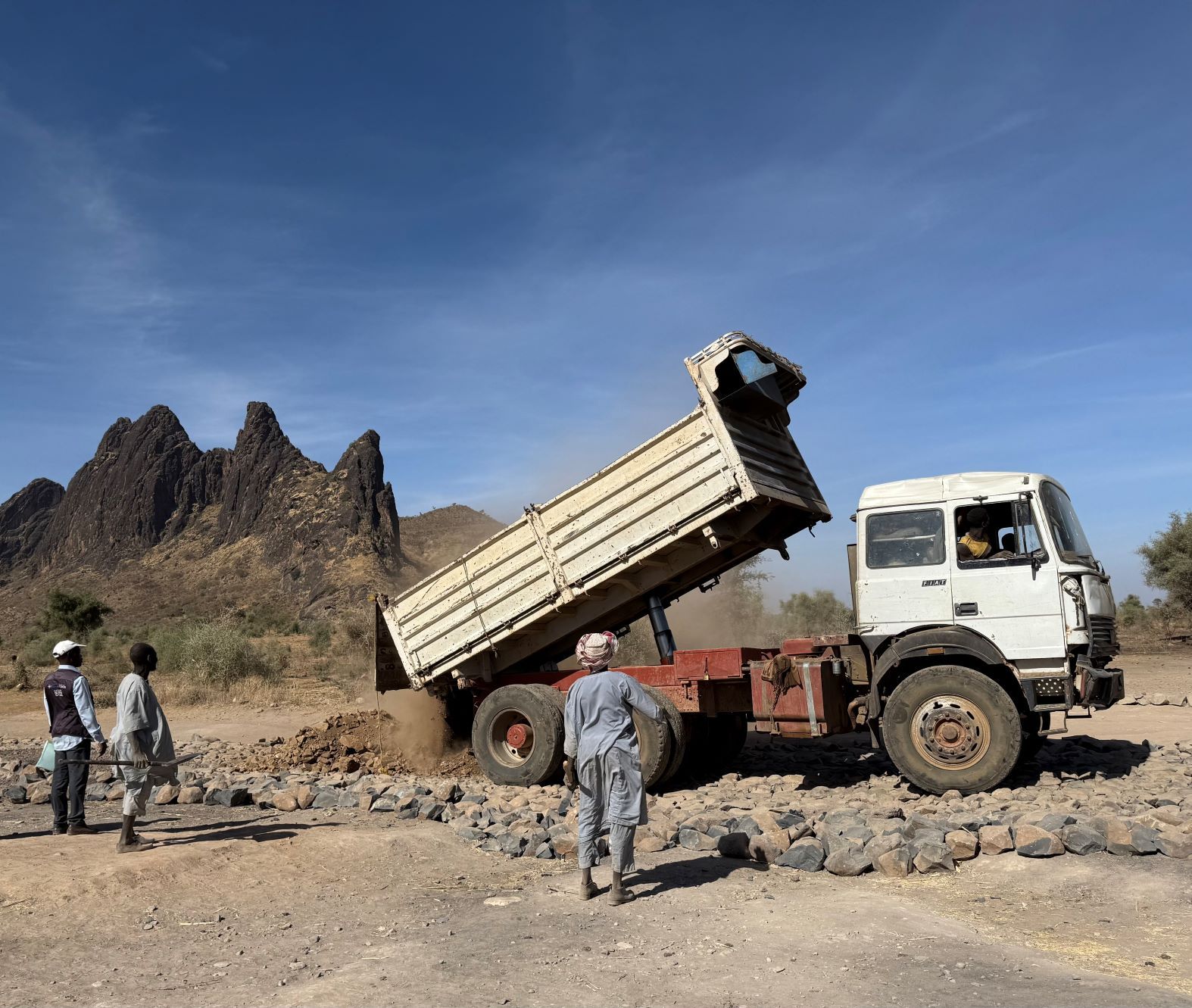 Tre uomini sono in piedi davanti a un camion che scarica terra per i lavori di riabilitazione della strada che porta a un'importante area di mercato nel Darfur.