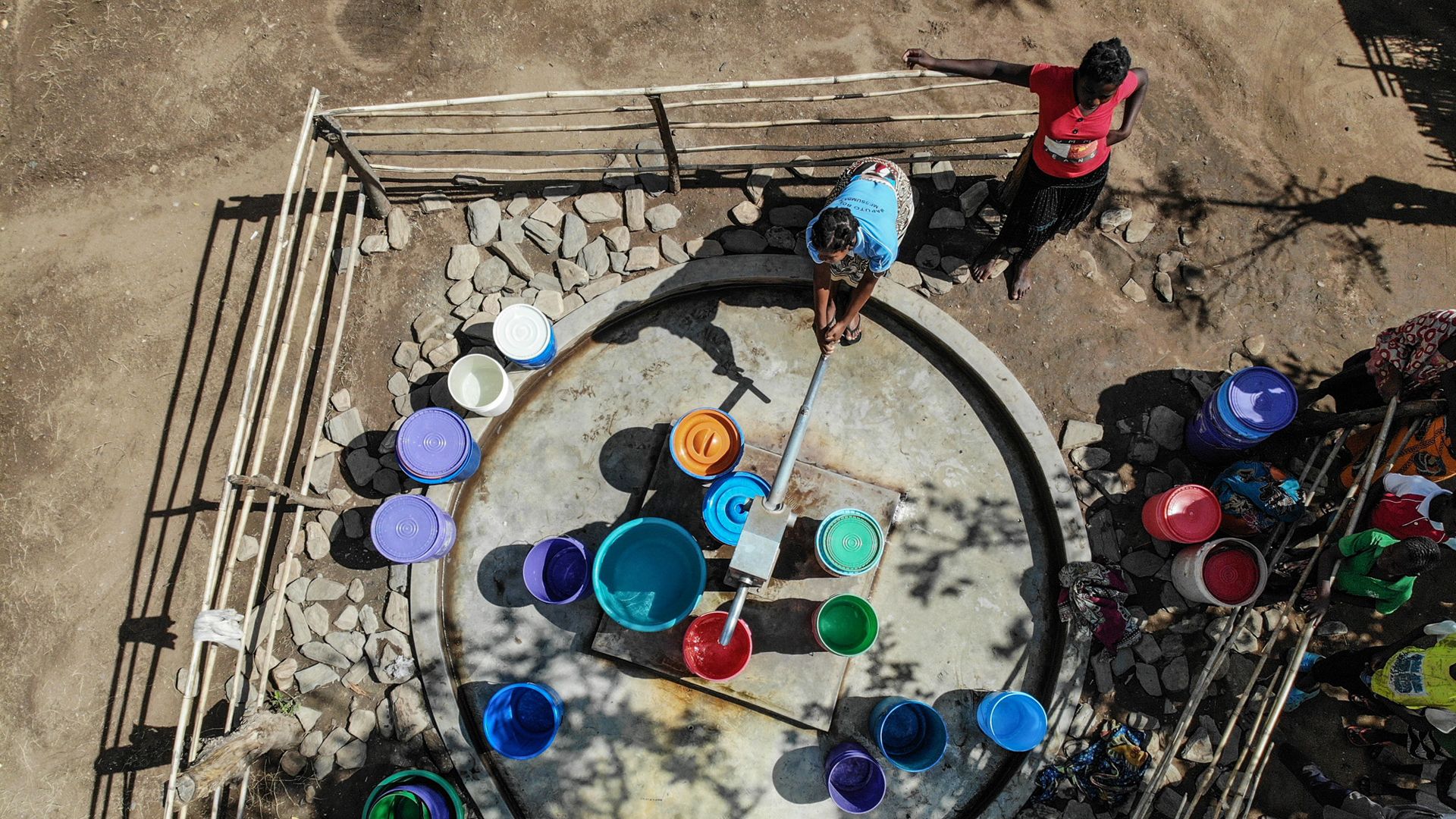 Una donna utilizza una pompa per estrarre acqua, circondata di secchi colorati già colmi, mentre un’altra donna la guarda.