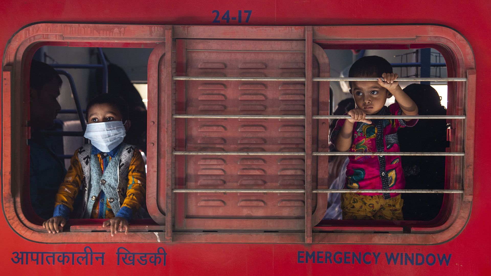 Due bambini guardano dalla finestra scorrevole di uno sportello: uno indossa la mascherina, l’altro no.