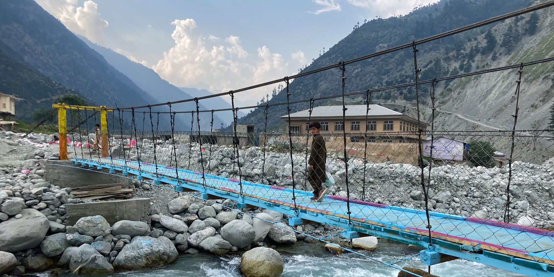 Un ragazzo attraversa un ponte di corda vicino a Mankiala, in Pakistan. Una distesa di grandi pietre costeggia un ruscello.