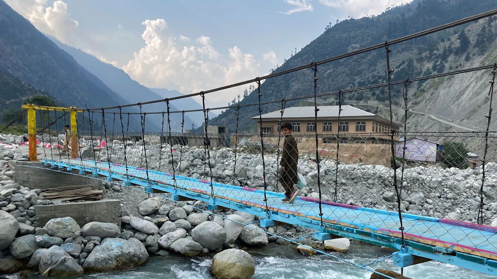 Un ragazzo attraversa un ponte di corda vicino a Mankiala, in Pakistan. Una distesa di grandi pietre costeggia un ruscello.