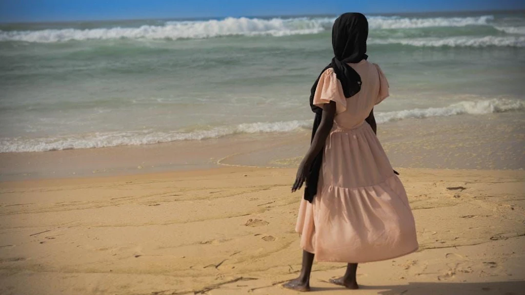 Una ragazza vestita con un abito color pastello è in piedi su una spiaggia e guarda il mare. La testa è coperta da un foulard nero.